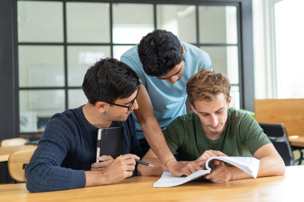 Serious students focused on class assignment. Three young men reading one textbook together in classroom. Studying and teamwork concept.